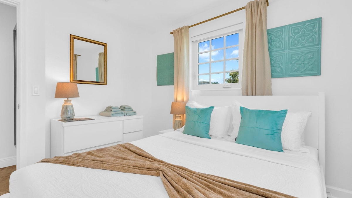 Este tercer dormitorio ventilado cuenta con una cama tamaño queen con sábanas blancas frescas, almohadas con detalles costeros y un televisor inteligente montado en la pared. Las ventanas luminosas aportan luz natural, creando un refugio tranquilo perfecto para relajarse después de un día explorando Palm Beach.