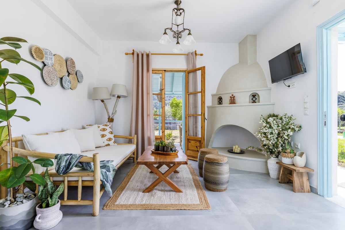 A comfortable seating area features a natural wood bench adorned with decorative pillows. A coffee table sits at the center, complemented by a textured rug. A stone fireplace with decorative items and two standing lamps are present, while light filters through a large window with sheer curtains.
