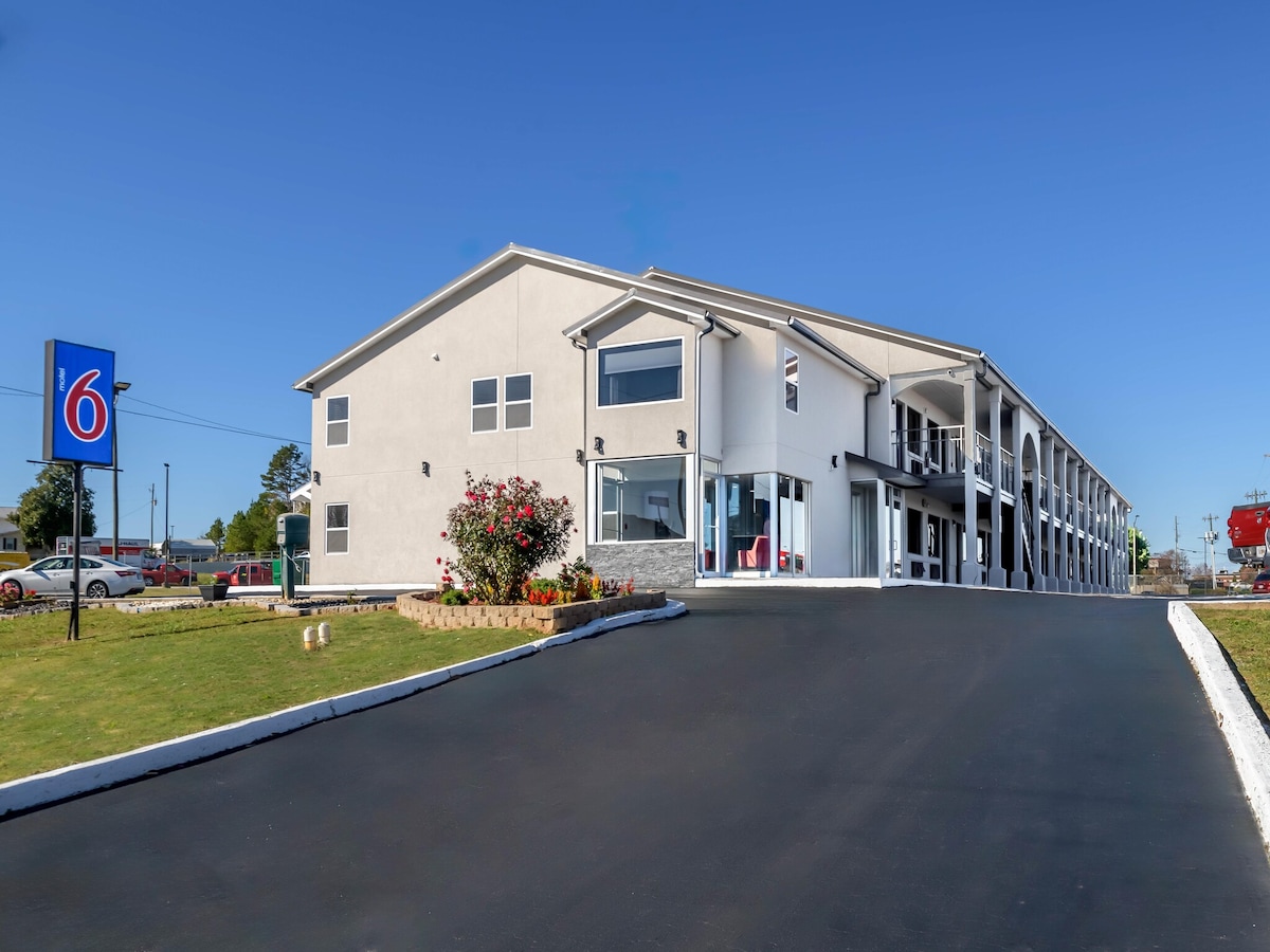 The exterior of the motel is visible, featuring a two-story building with large windows. A manicured lawn with landscaped flower beds is seen in the foreground. The driveway is paved, leading up to the entrance, located beneath a clear blue sky.