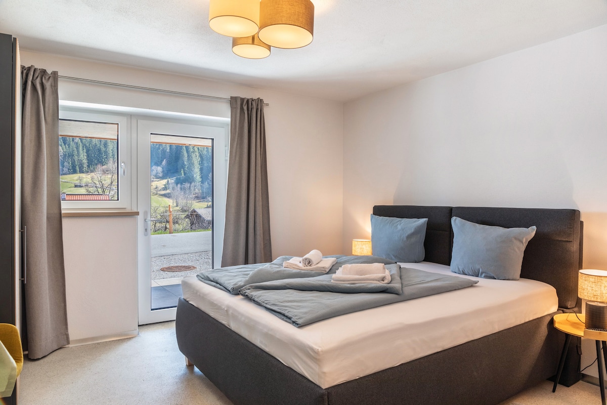 A spacious bedroom features a comfortable bed with soft linens and decorative pillows. Large windows bring natural light into the room, revealing views of the surrounding landscape. Two bedside lamps provide additional illumination, while gray curtains frame the windows.