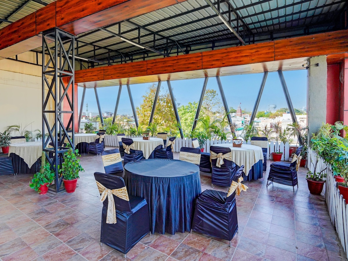 An outdoor dining area is presented, featuring round and rectangular tables covered with white tablecloths and dark blue skirts. Chairs with gold sashes complement the tables. Large windows provide views of greenery and the surrounding city, enhancing the pleasant atmosphere.