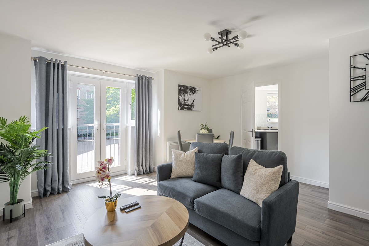 A modern living area features a plush gray sofa with decorative pillows facing a round wooden coffee table. Natural light streams in through double doors leading to a balcony, while a dining area with a table is visible in the background. Potted plants add a touch of greenery.