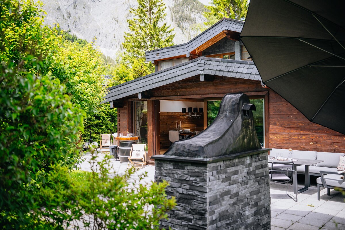 The exterior of a modern chalet is framed by lush greenery, showcasing sleek wooden architecture. A stylish stone feature is positioned in the foreground. Outdoor seating is arranged under a shaded area, providing a comfortable space for relaxation.