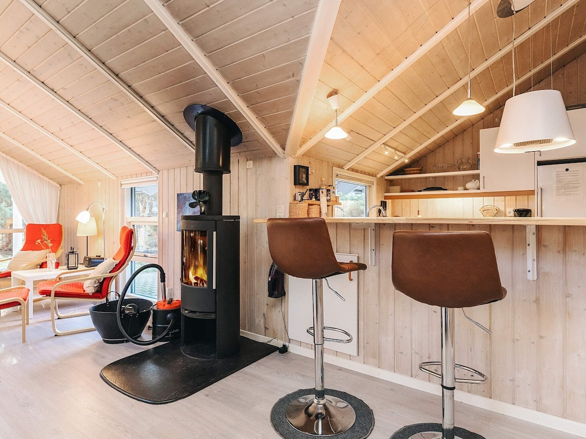 An inviting interior features a cozy fireplace surrounded by wooden walls. The bar area includes two stylish stools, offering a casual dining space. Natural light filters through the windows, highlighting the warmth of the overall ambiance.