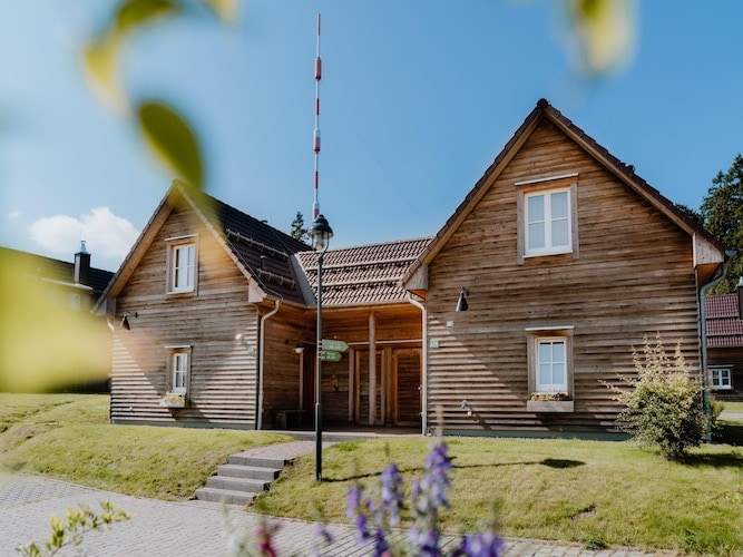 A charming wooden holiday home is displayed, featuring a symmetrical design with two gabled sections. Light-colored wooden siding and white window frames contribute to a rustic aesthetic. A well-maintained lawn and flower beds surround the entrance, offering a serene atmosphere against a clear blue sky.
