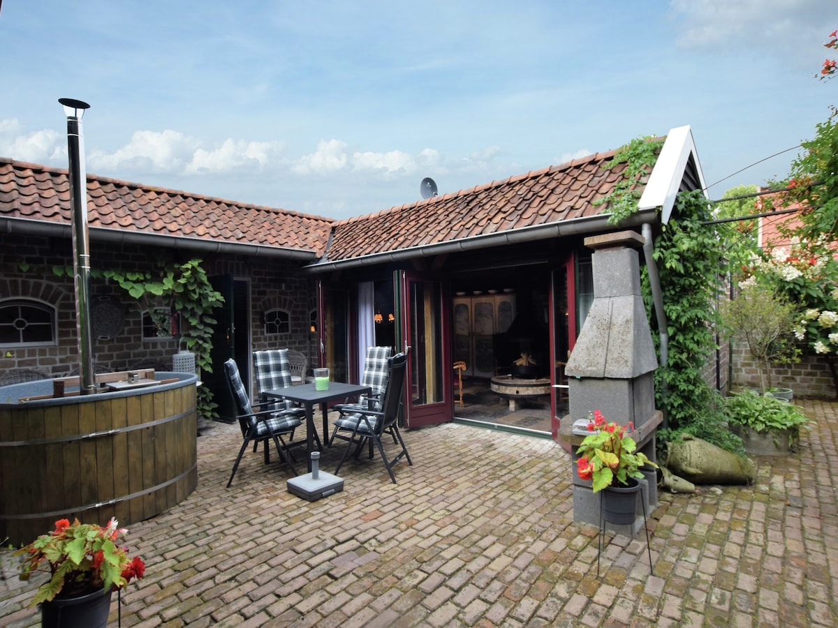 An outdoor terrace is surrounded by greenery and features a wooden hot tub, dining table with chairs, and flower pots. The adjoining building showcases large glass doors that provide a glimpse into the cozy living area. The brick patio adds a rustic charm to the setting.