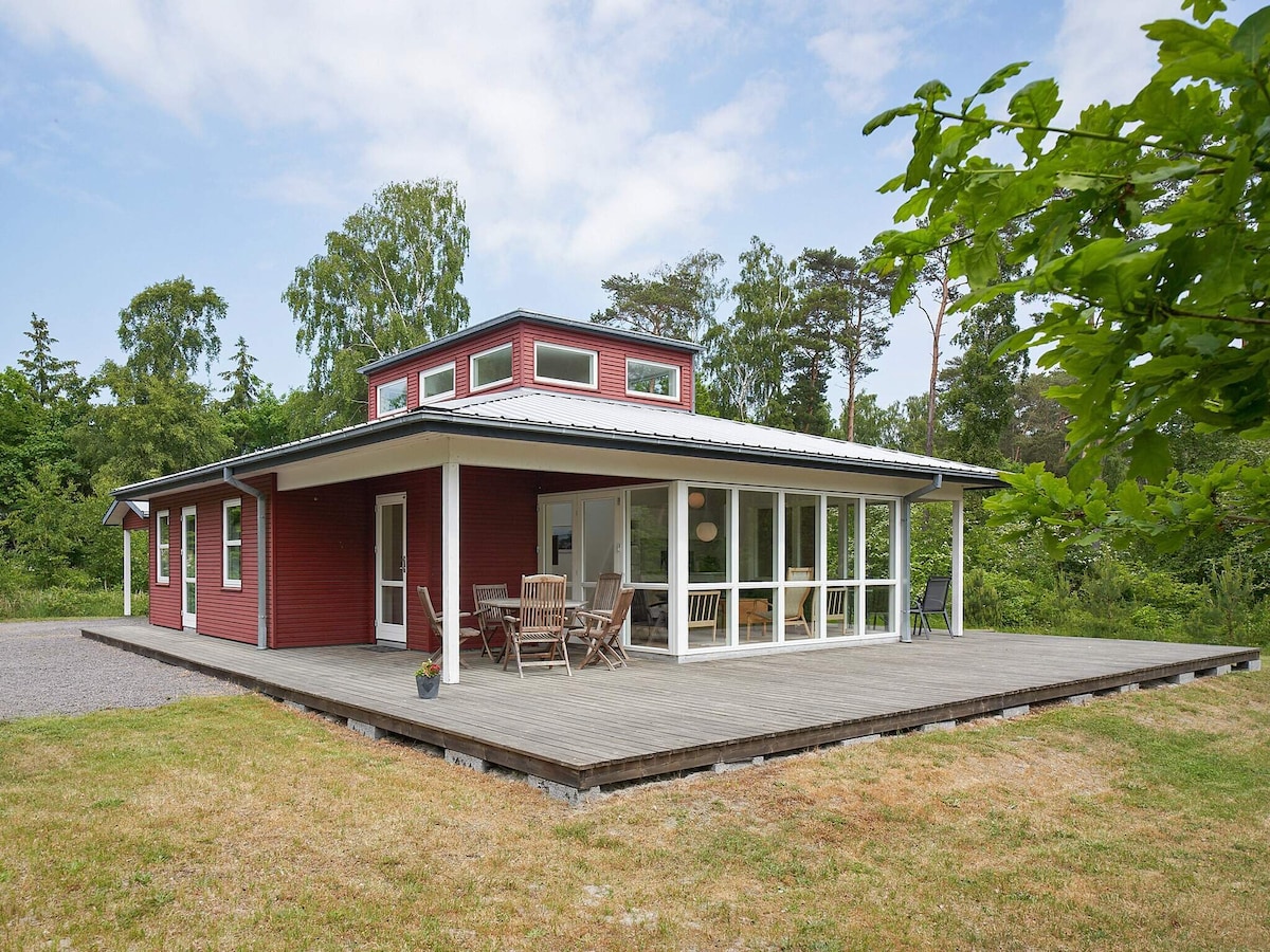 A spacious holiday home is set amidst greenery, featuring a large wooden deck and multiple glass doors. Outdoor seating is provided for dining, with a relaxed atmosphere inviting enjoyment of the natural surroundings.