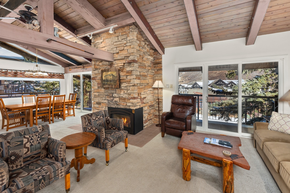 A spacious living area is highlighted by a stone fireplace and vaulted wooden ceilings. Comfortable seating includes patterned armchairs and a sectional sofa, arranged around a rustic wooden coffee table. Large windows frame the view of the surrounding landscape, allowing natural light to fill the space.