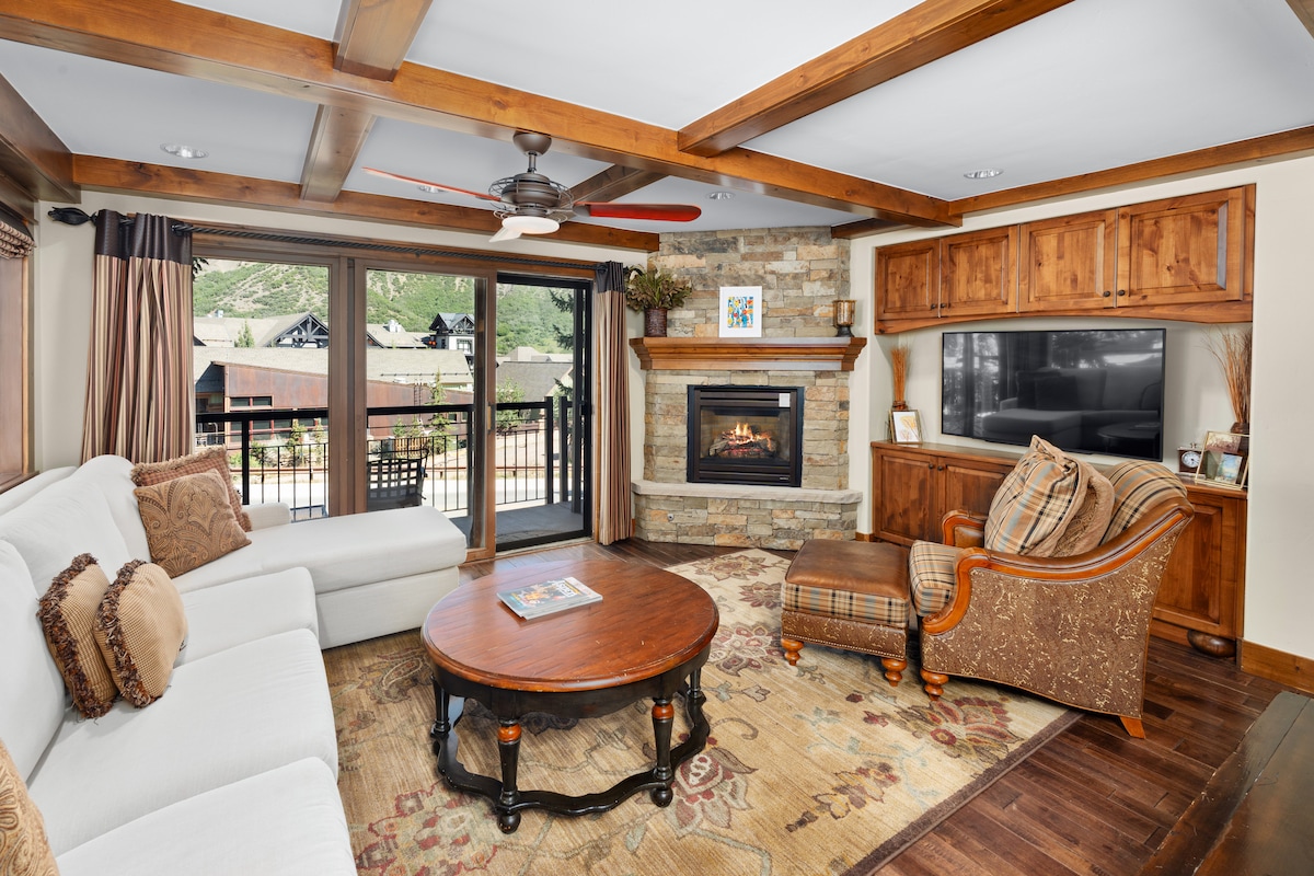 A cozy living area features a white sectional sofa and a richly colored area rug. A circular coffee table sits at the center, while a stone fireplace adds warmth. Natural light streams in through large windows, showcasing mountain views and enhancing the inviting ambience.