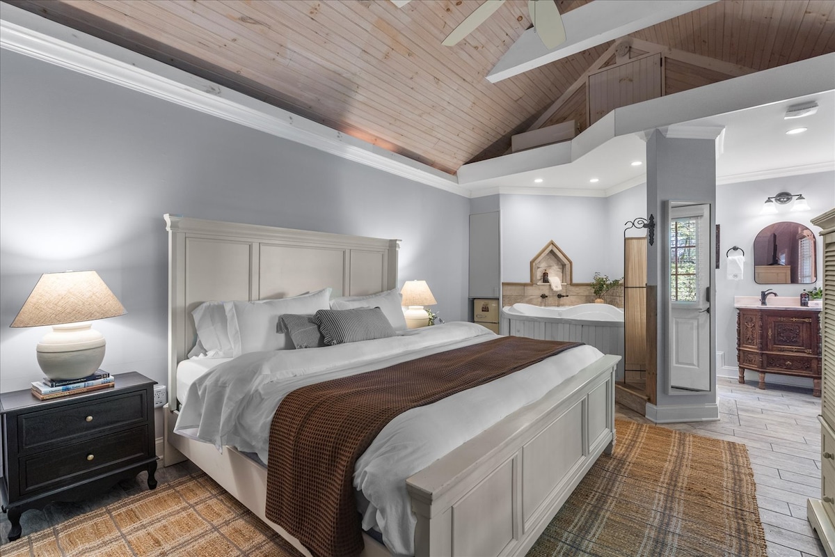 A spacious bedroom features a comfortable king-sized bed dressed in crisp linens. Soft lighting is provided by bedside lamps on dark wood nightstands. To one side, a jetted tub is partially visible, complementing the warm wooden accents of the ceiling and flooring.