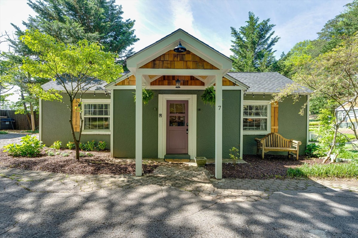 A charming cottage exterior is framed by mature trees and landscaped gardens. The inviting entrance features a light purple door and a covered porch. A wooden bench sits beside the path, complementing the natural surroundings. Soft sunlight illuminates the facade, creating a welcoming atmosphere.