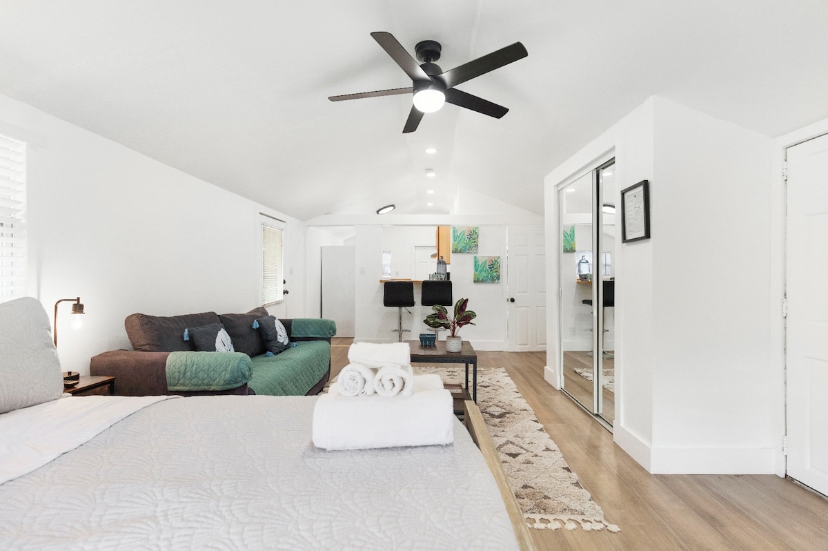 An open living area is highlighted by a spacious bed with neatly arranged towels nearby. A comfortable sofa and a vibrant green accent chair are present, alongside a coffee table. Light hardwood flooring complements the bright walls, while a ceiling fan provides air circulation.