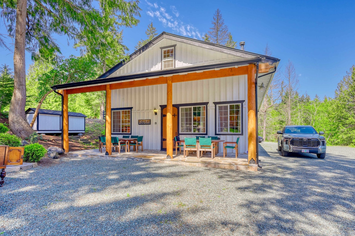 The exterior of the cabin features exposed wooden beams and a welcoming porch with several green chairs arranged around a rustic wooden table. A gravel driveway offers parking for vehicles, and lush greenery surrounds the property, providing a serene setting.