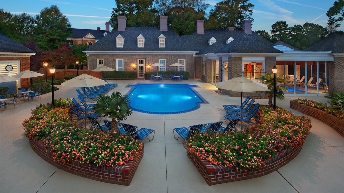 An outdoor pool area is surrounded by well-maintained gardens featuring vibrant flowers and palm trees. Lounge chairs are arranged neatly by the pool, and shaded areas with umbrellas offer relaxation spots. The charming brick building in the background complements the serene atmosphere.