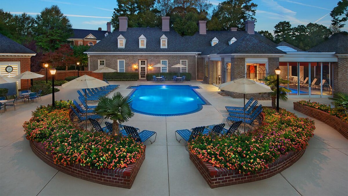 An outdoor pool area is surrounded by vibrant flower beds and lounge chairs. Sun umbrellas offer shade beside the clear pool, and a brick wall adds structure to the garden. The backdrop features a stately building with a shingled roof and large windows, creating a relaxing atmosphere.