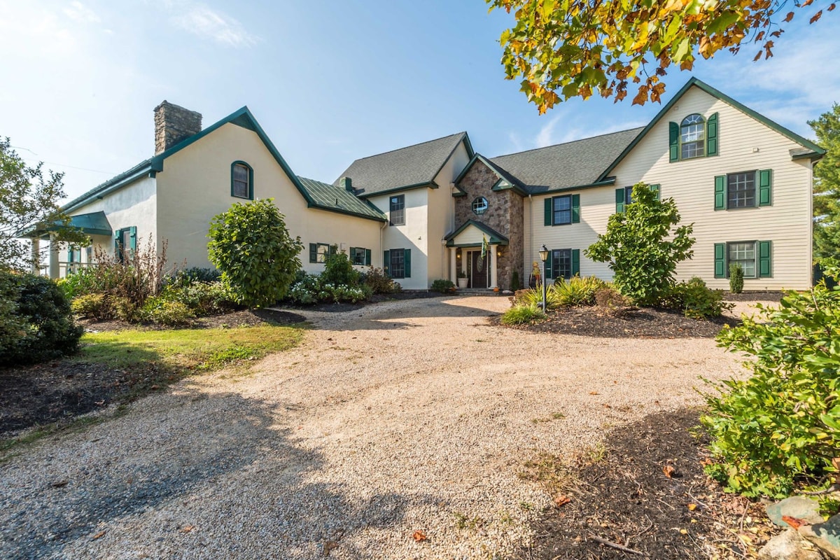 The exterior of the estate is presented, showcasing a spacious façade with a combination of light-colored stucco and dark green accents. The surrounding landscape features a well-maintained garden with shrubs and trees, along with a gravel driveway leading to the entrance.