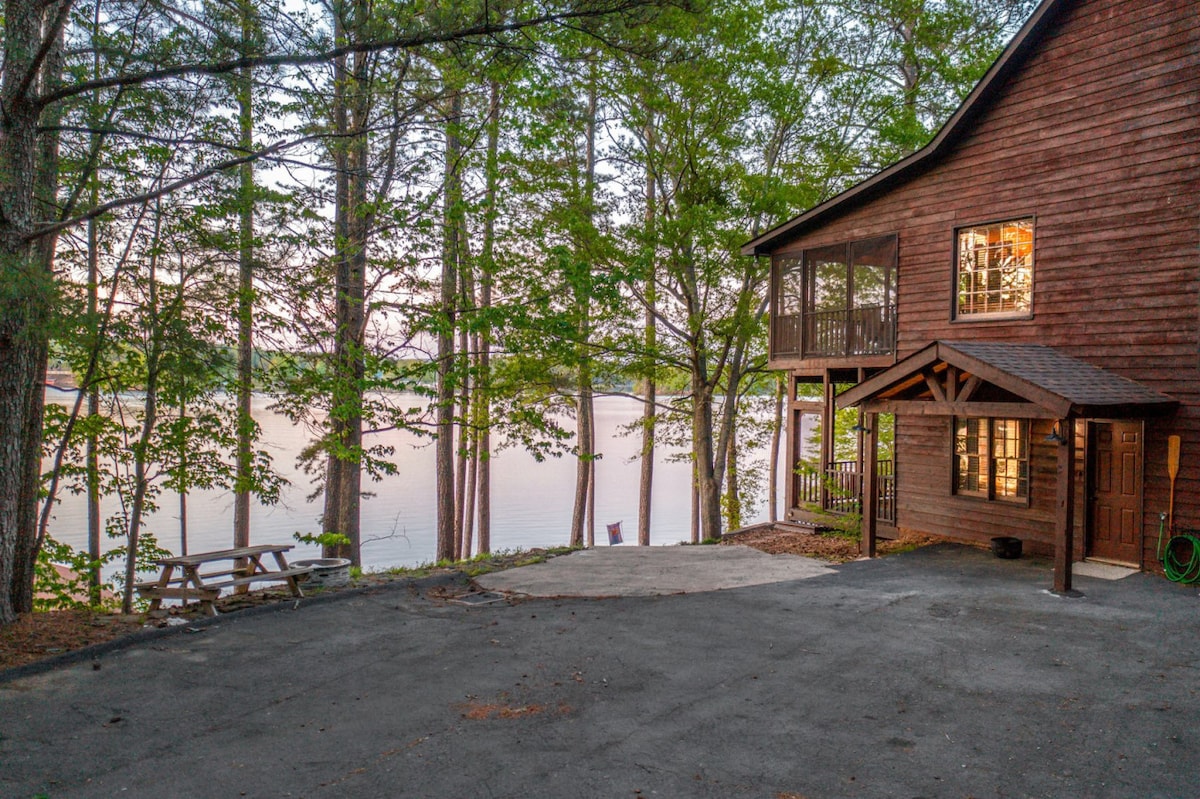 The exterior of the home is framed by tall trees, providing a natural setting alongside the water. A gravel driveway leads to the entrance, while a wooden picnic table is positioned nearby, facing the serene lake view. Light reflects off the water in the background.