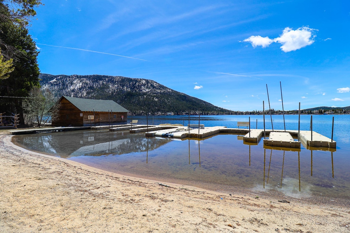 A serene view of a sandy beach area extends to the calm waters of Grand Lake. Wooden boat docks are visible in the foreground, with a rustic cabin situated nearby against a backdrop of mountains and a clear blue sky.