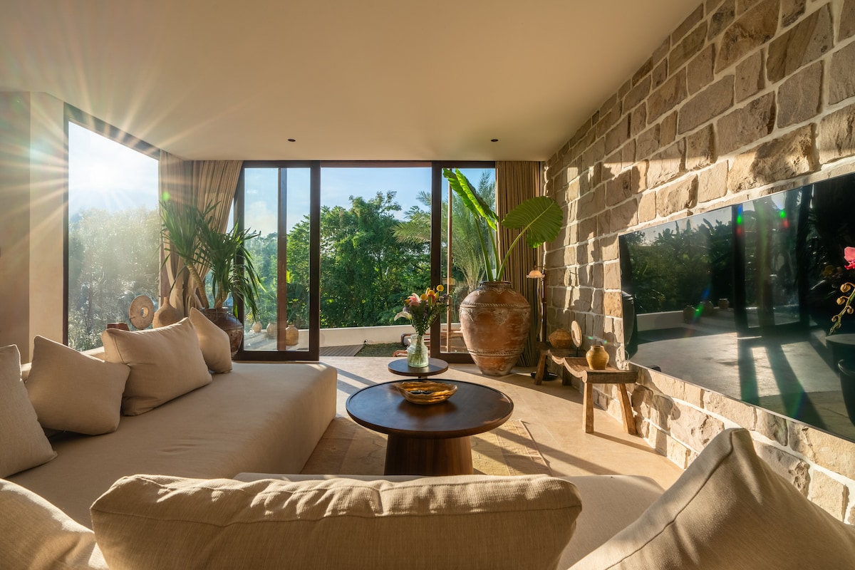 A spacious living area is illuminated by natural light, highlighting stone walls and large windows that open to lush greenery. A comfortable sofa is paired with a round coffee table, adorned with decorative items and plants, creating a serene and inviting atmosphere.