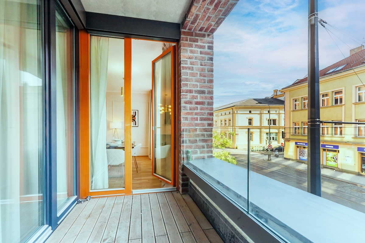 A balcony space features wooden flooring and glass railings, offering an unobstructed view of the street below. The facade displays a combination of modern and traditional architecture, with large glass doors opening to the interior living area.
