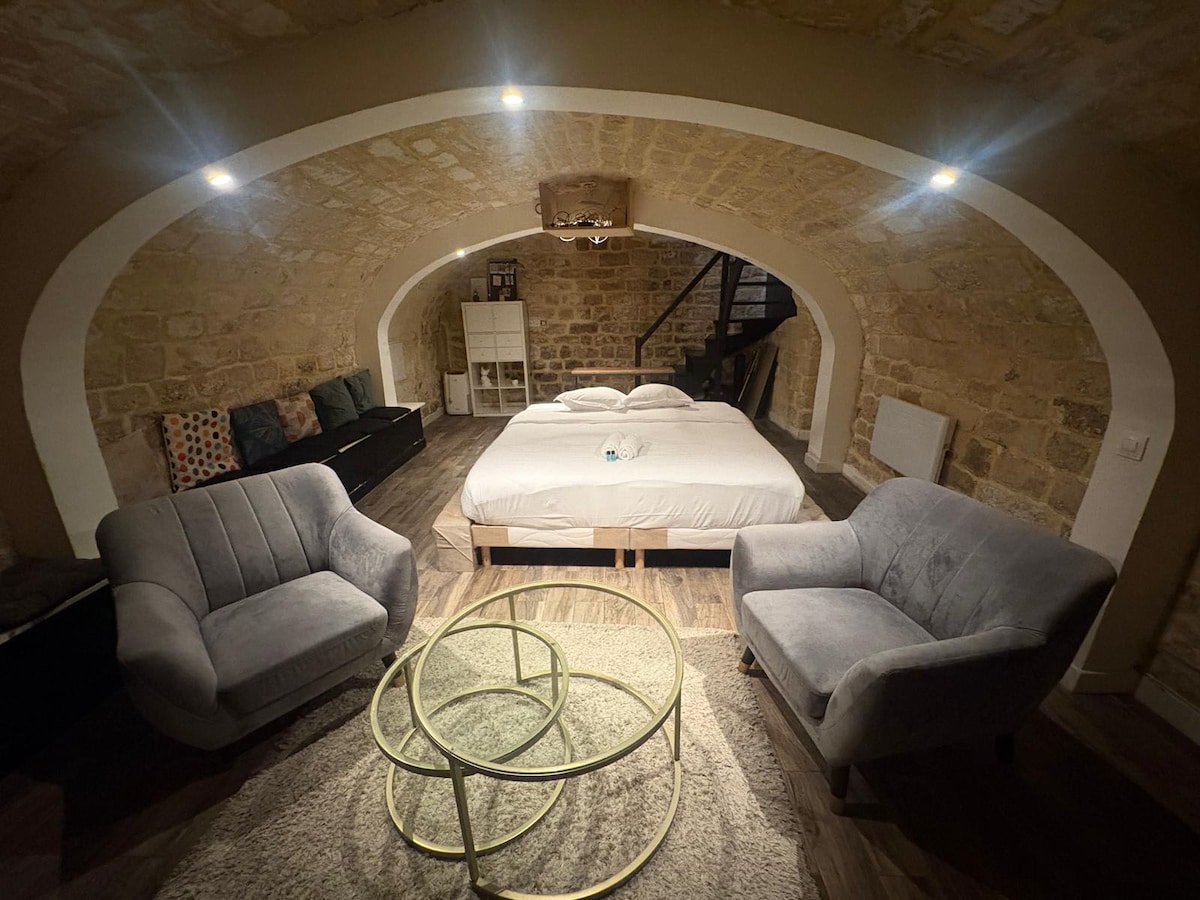 An inviting studio space features a spacious bed situated beneath an arched stone ceiling. Two comfortable armchairs flank a circular coffee table on a soft area rug. Subtle lighting highlights the stone walls, creating a cozy ambiance in this well-designed environment.