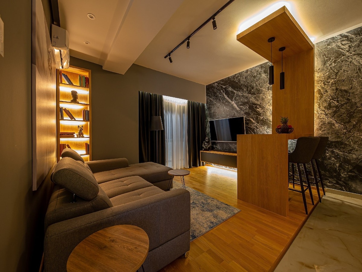 The spacious living area features a modern grey sofa positioned next to a small round table. A flat-screen TV is mounted on a dark feature wall, while warm accent lighting illuminates a nearby bookshelf. Large windows are dressed with dark curtains, allowing soft natural light into the space.