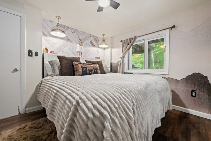 Cozy chalet-style bedroom with plush king bedding, soft lighting, forest views, and warm mountain-inspired decor. Peaceful retreat designed for restful nights after hiking, skiing, or exploring the White Mountains. and ceiling fan, full length mirror