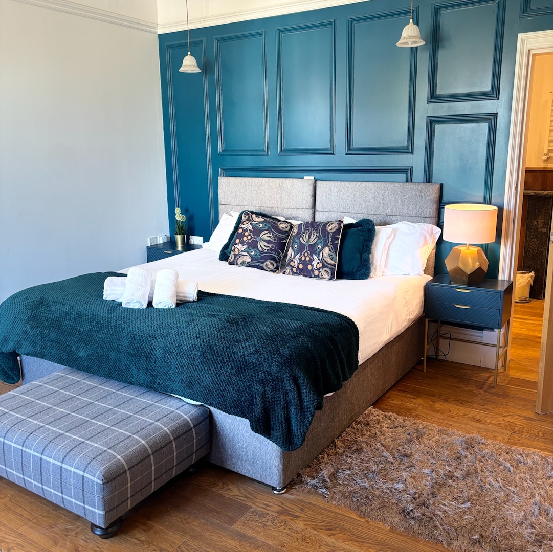 A spacious bedroom features a comfortable bed with plush pillows and a soft, textured blanket. Two pendant lights hang above nightstands with lamps, while a decorative ottoman adds a touch of style. Light wooden flooring and a cozy rug create a welcoming ambiance.