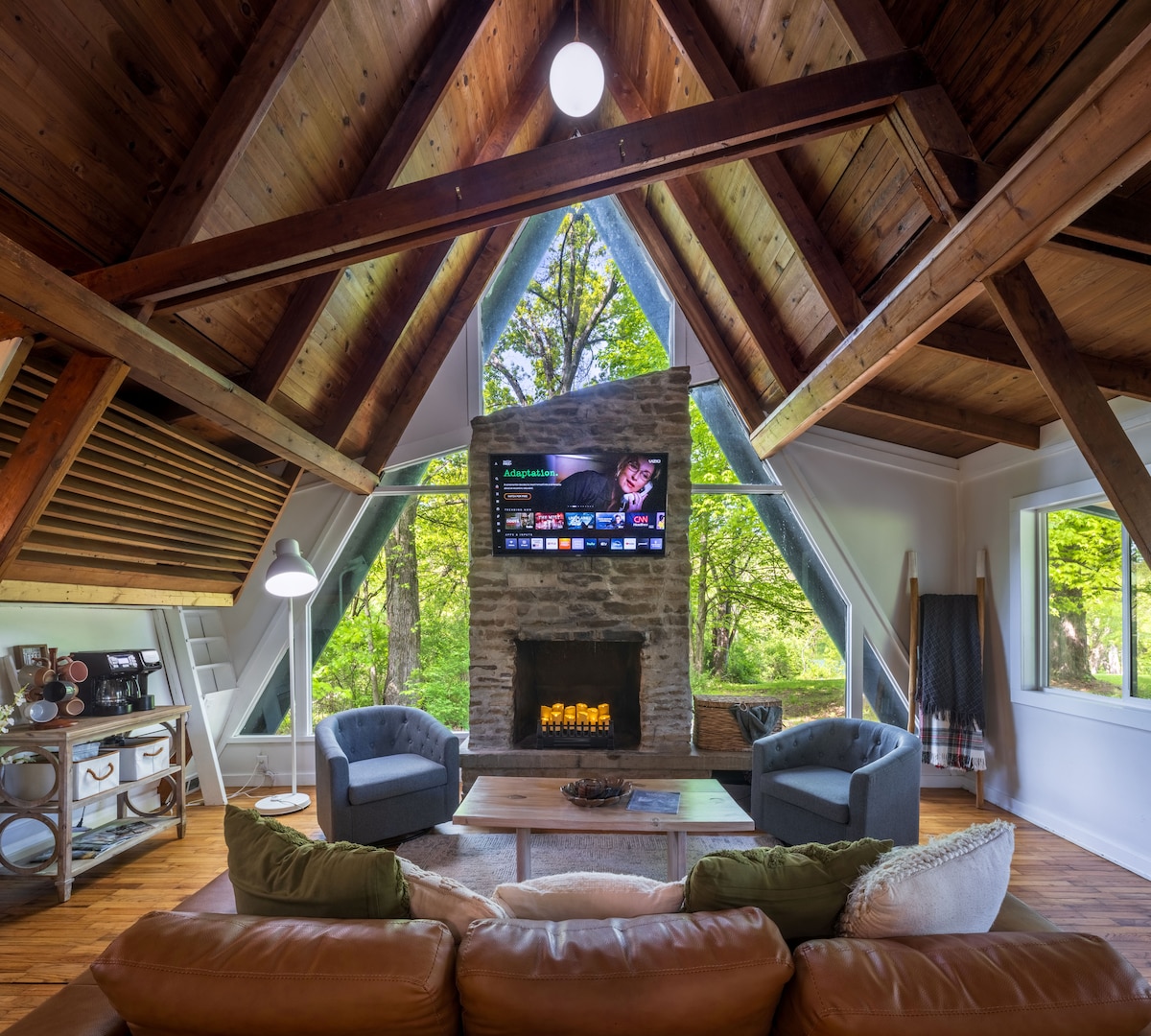 The living area is characterized by high cathedral ceilings and large windows, allowing natural light to illuminate the space. A stone fireplace serves as a focal point, while a smart TV is mounted above it. Comfortable seating includes a brown couch and two armchairs, set against a backdrop of lush greenery outside.