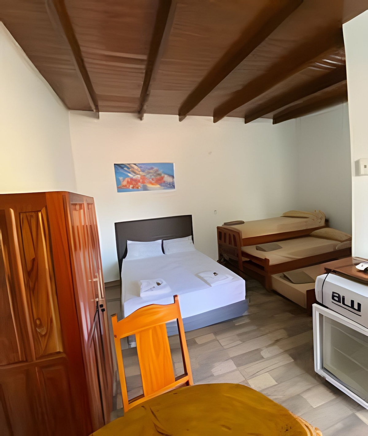 A cozy sleeping area is visible, featuring a spacious bed and a single bed. Wooden beams accent the ceiling, while a simple dresser and a small refrigerator are present. A round table with a chair offers a functional space, and a wall art piece adds a splash of color.