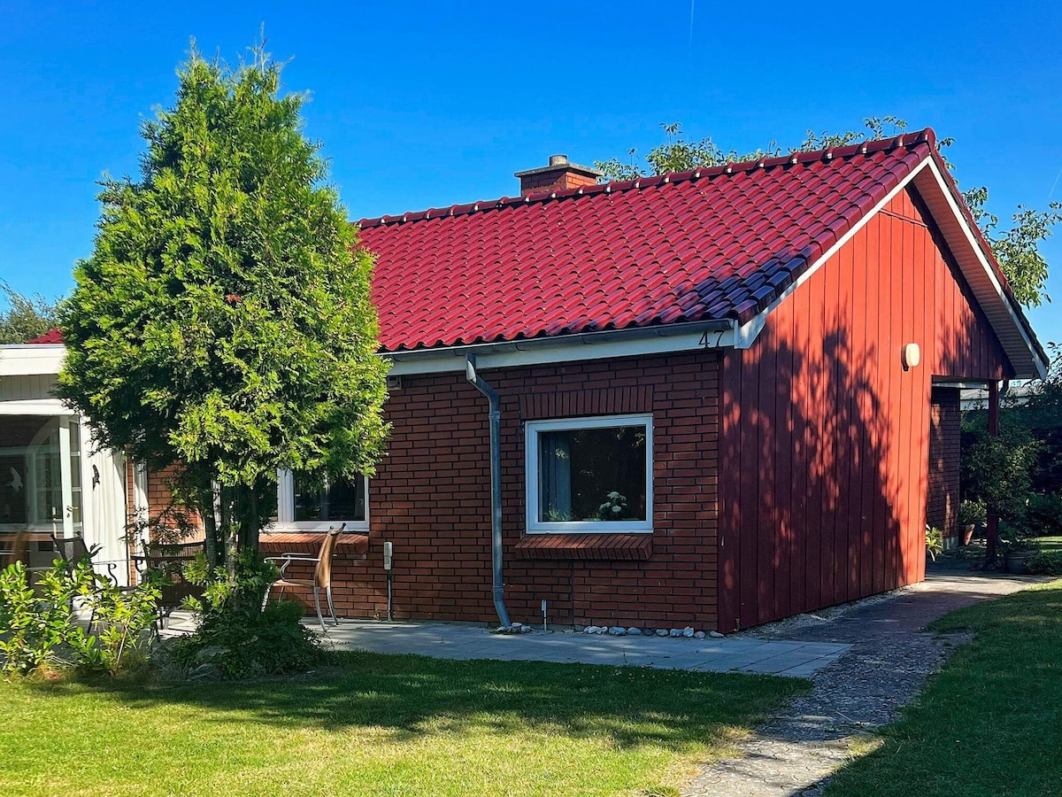 The holiday home features a brick exterior with a vibrant red roof. A well-maintained garden surrounds the building, complemented by a small tree to the left. Large windows allow natural light to enter, enhancing the inviting appearance of the property.