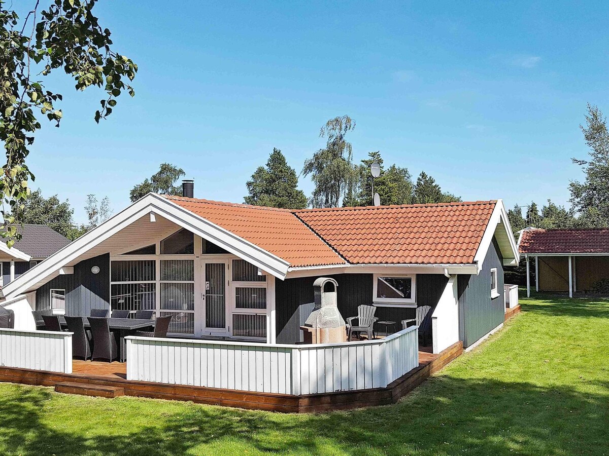 A holiday home with a sloped roof and a red-tiled covering is situated on a grassy area. The exterior features large windows for natural light. A wooden terrace surrounds the house, accompanied by outdoor seating and a barbecue for guests to enjoy.