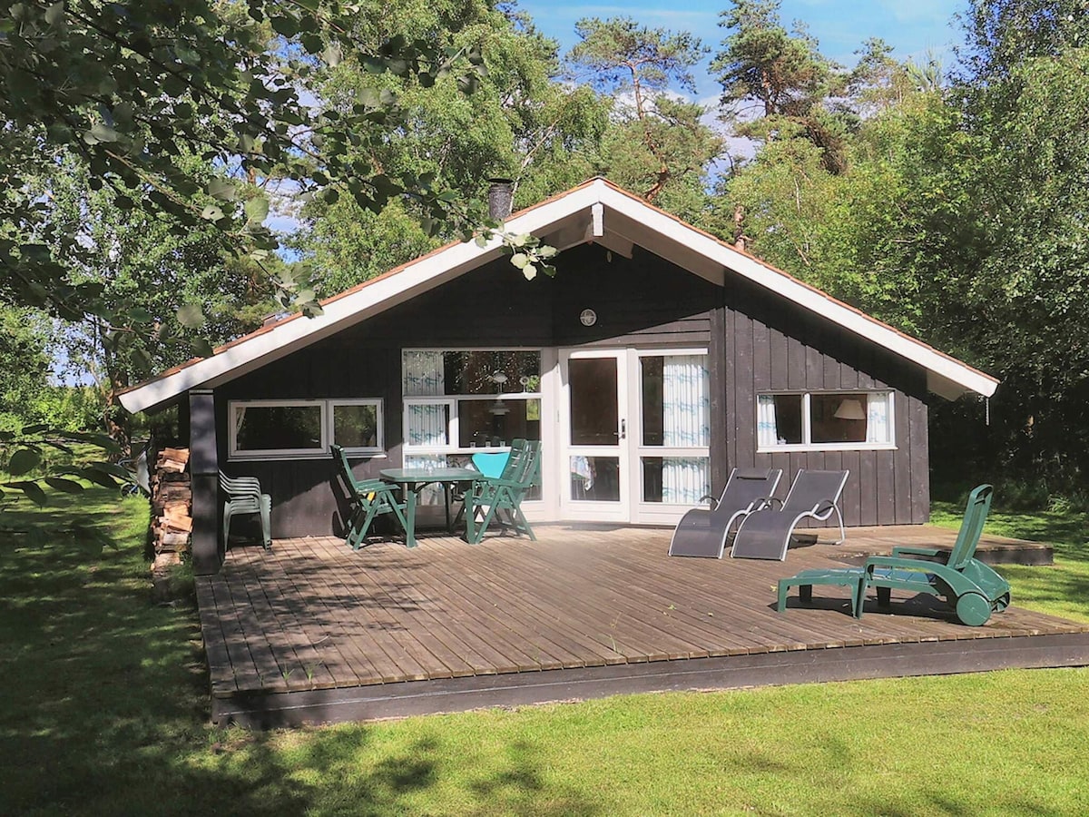 A holiday home is presented with a sloping roof and wooden exterior, situated in a lush green setting. Two lounge chairs and a dining table with chairs occupy the spacious wooden deck, inviting relaxation. Large glass doors provide access to the interior, allowing natural light to fill the space.