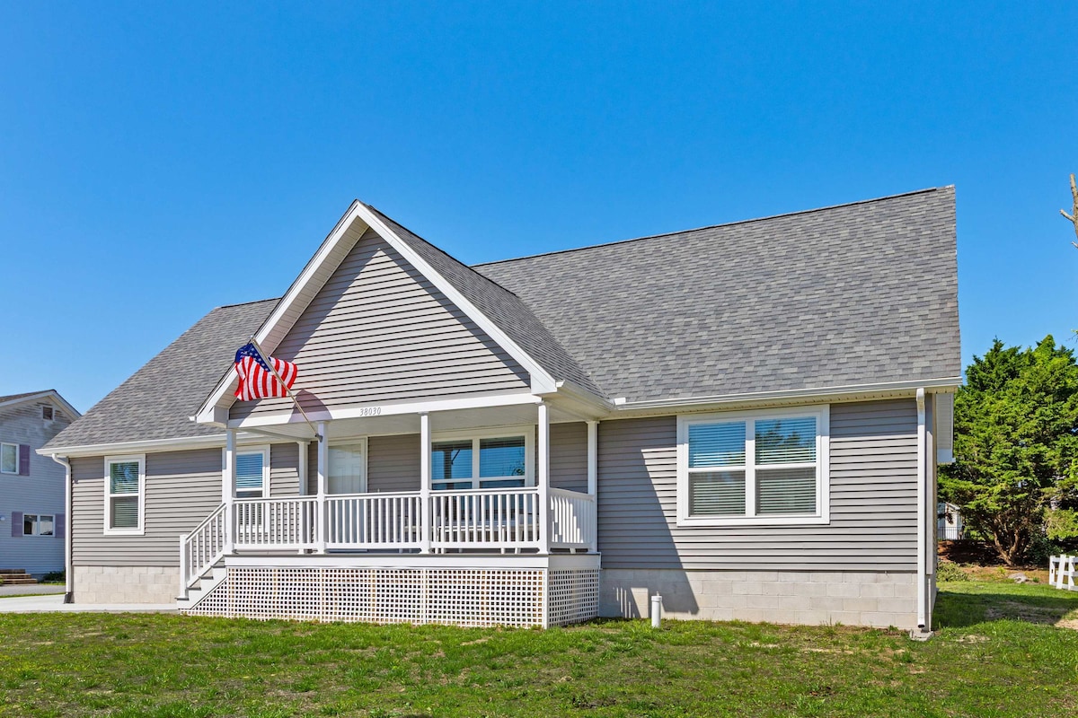 A spacious single-story beach house is framed by a clear blue sky. The exterior features light grey siding and a welcoming front porch with white railings. An American flag hangs proudly above the entrance, and large windows allow natural light to flood the interior.