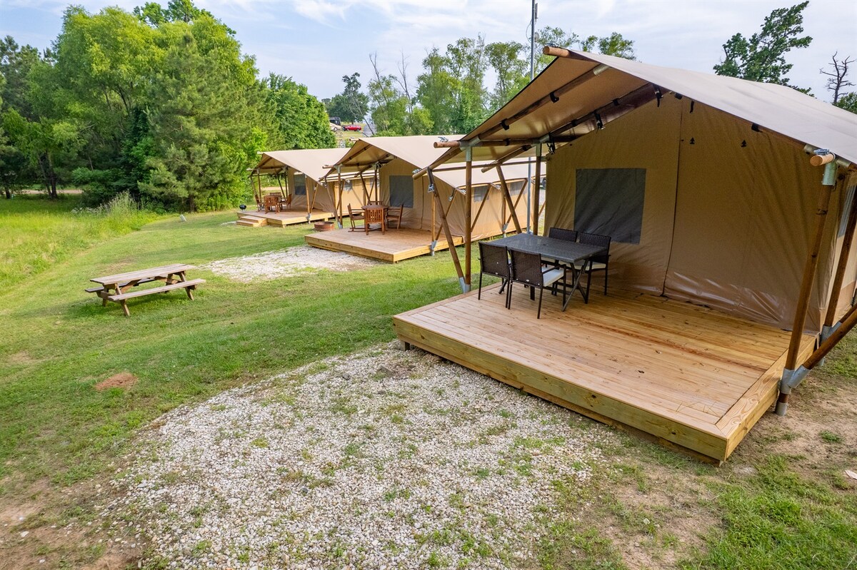 Several luxury glamping tents are set on raised wooden platforms, surrounded by natural greenery. Each tent features a covered porch area with seating options, and a picnic table is positioned nearby on a gravel surface. The tranquil outdoor setting invites guests to relax in nature.