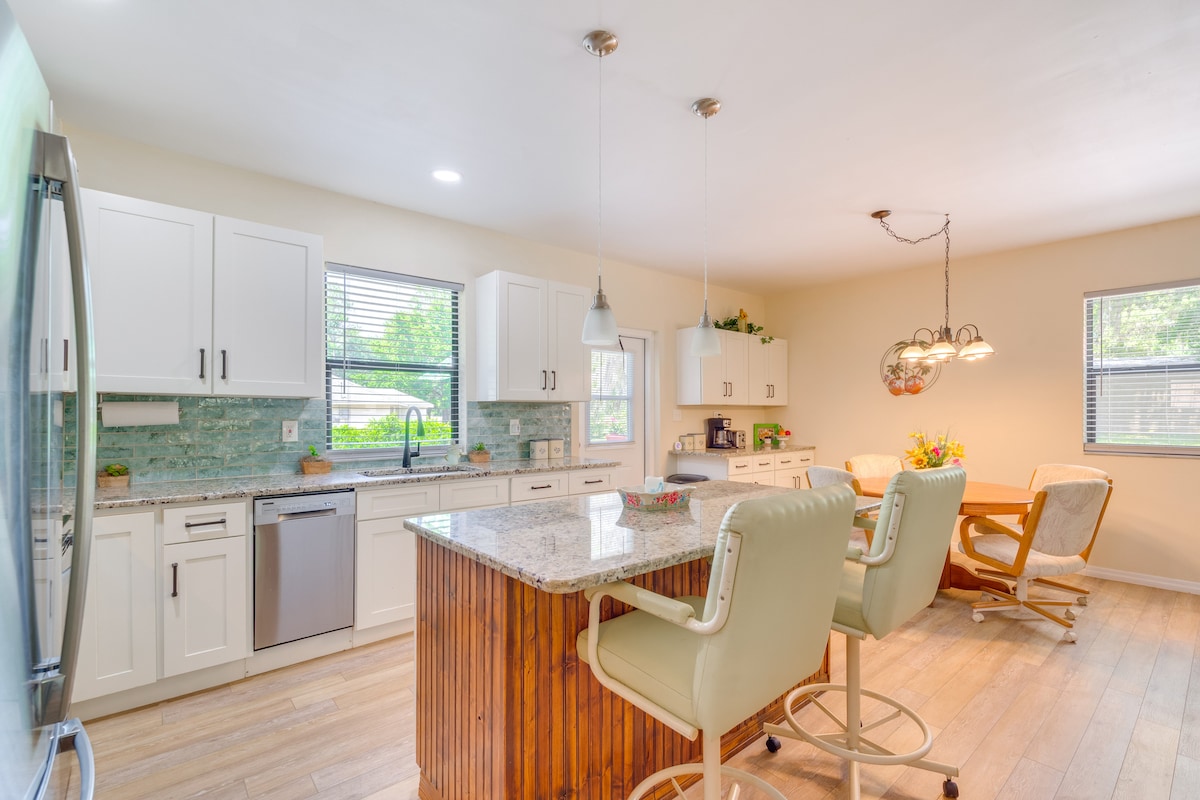 The modern kitchen features white cabinetry and a large island with a granite countertop. Seating is provided at the island, complemented by light-colored bar stools. An adjacent dining area includes a table surrounded by chairs, illuminated by natural light from the windows.