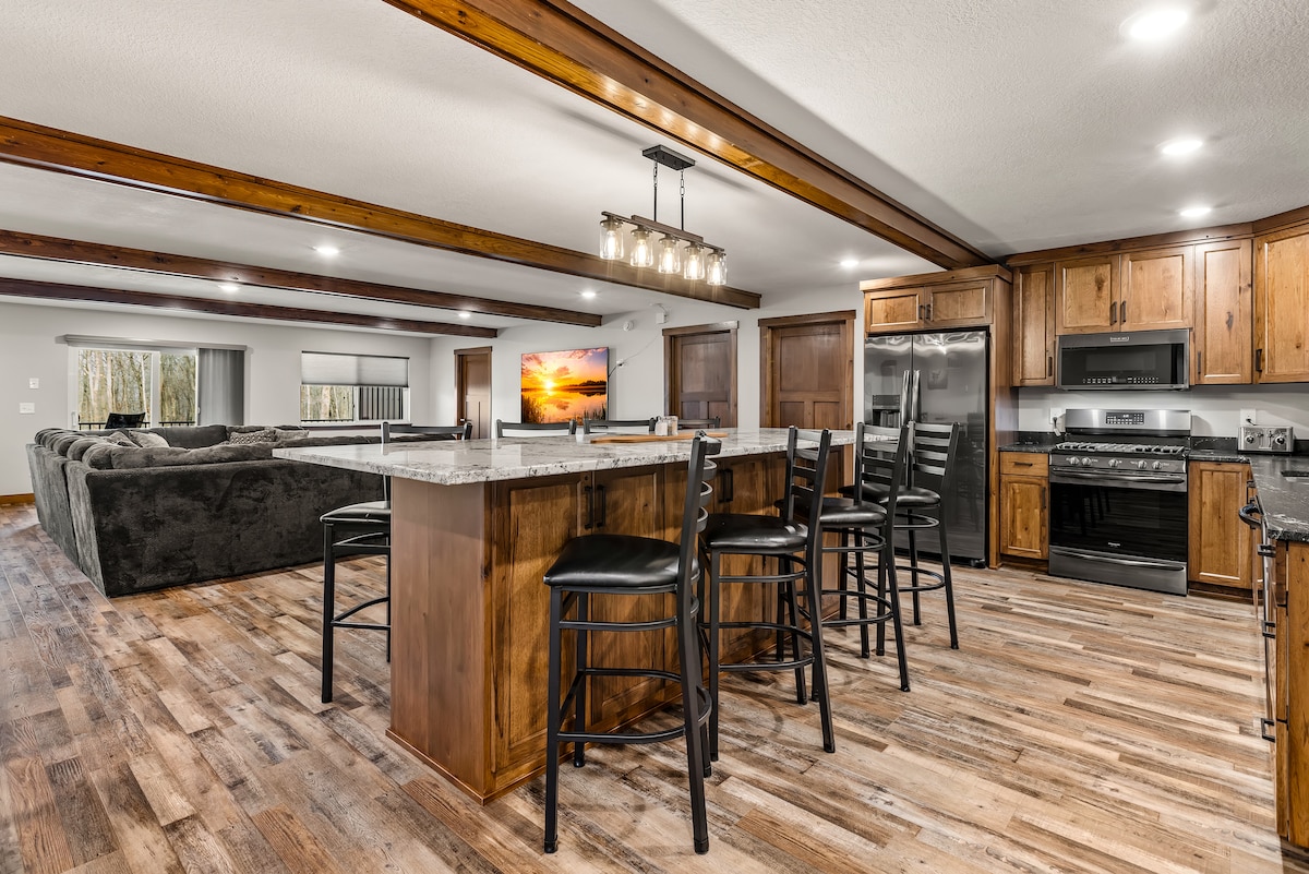 A spacious kitchen and dining area features a large island with seating for six, complemented by a modern light fixture. Natural wood cabinetry and stainless steel appliances enhance the inviting atmosphere. An open concept layout connects to a comfortable living area with plush seating and warm wood accents.