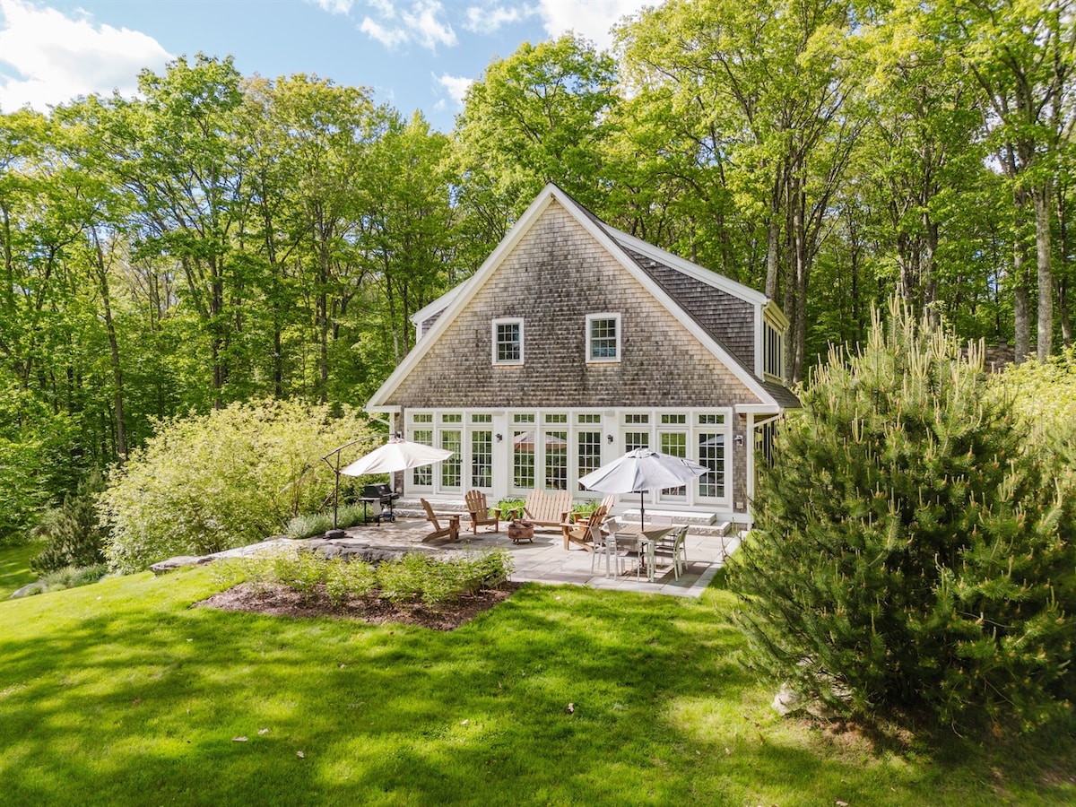 The guesthouse exterior features a charming two-story design with large windows. A spacious patio, surrounded by greenery, includes multiple Adirondack chairs and umbrellas, ideal for outdoor relaxation. Lush lawn areas complement the natural setting, providing a serene atmosphere amidst the trees.