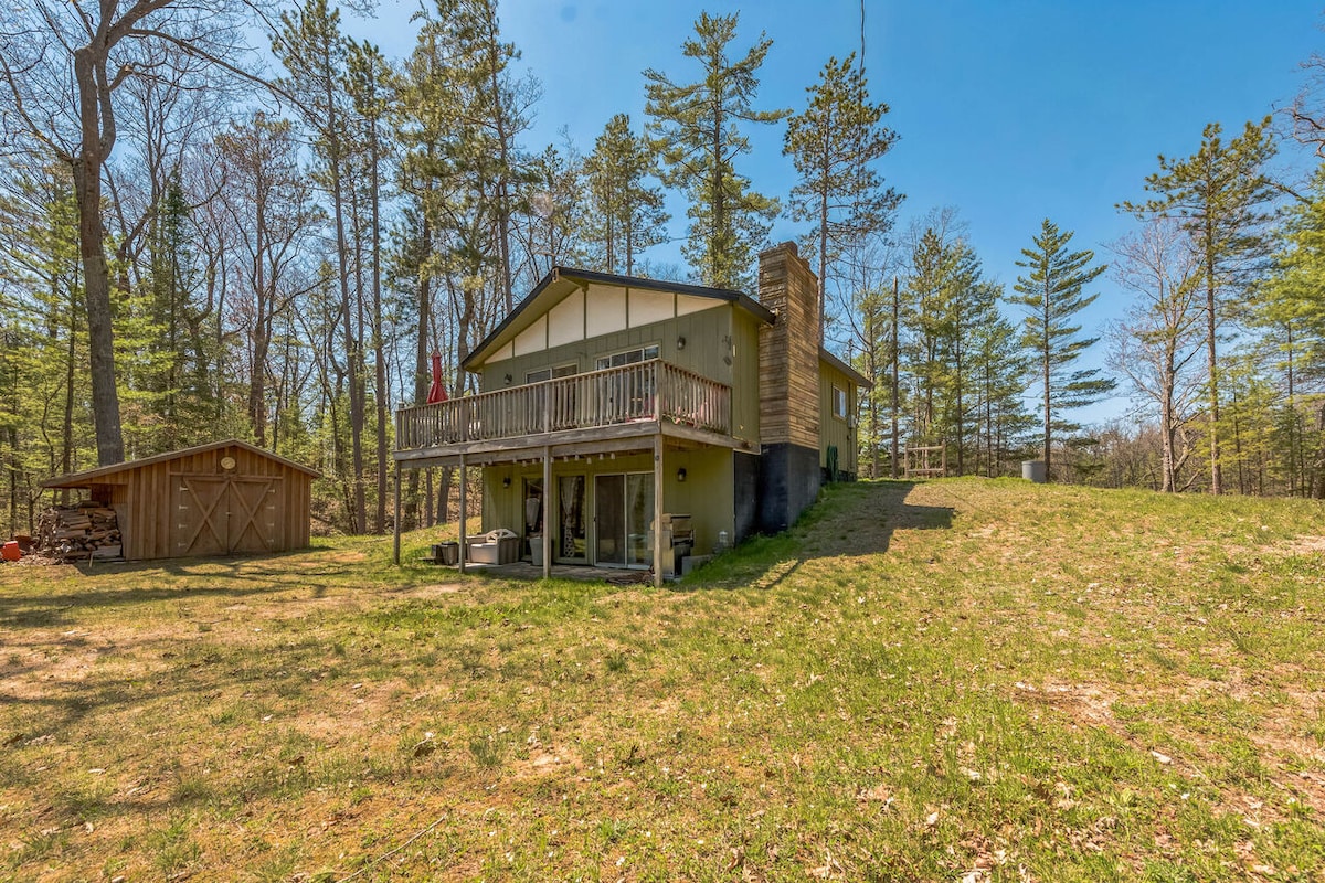 A spacious two-story cottage is set against a backdrop of tall trees. The exterior features a wooden deck with railing and a stone chimney, while a shed is positioned on the left. A grassy area extends from the house, providing a serene outdoor space.