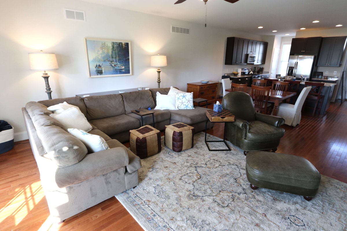 A cozy living area features a large sectional sofa and an armchair, complemented by a rustic wooden side table. Light-colored walls create a bright atmosphere, while a patterned area rug adds warmth beneath. The space includes a dining area and a view of the kitchen in the background.