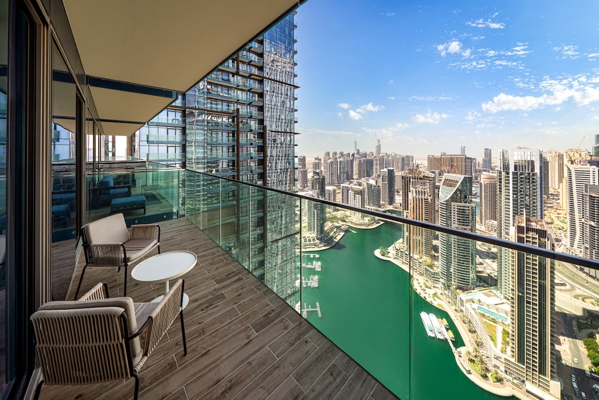 An outdoor balcony is showcased, featuring two modern chairs and a small round table. A panoramic view of Dubai Marina and the city skyline is present, with glistening water below and high-rise buildings stretching into the distance under a clear blue sky.