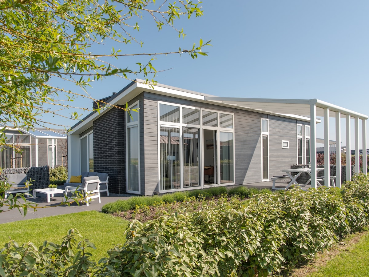 A modern chalet is situated within lush greenery, featuring large glass windows that invite natural light into the interior. A covered terrace with seating enhances the outdoor space, and well-maintained shrubs line the pathway, providing a serene atmosphere.