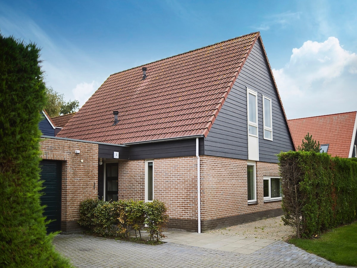 The exterior of a detached accommodation is presented, featuring a mix of textured brick and dark paneling. A sloped roof adds character, and well-maintained shrubs frame the entrance. A paved pathway leads to the door, enhancing accessibility to the welcoming space.