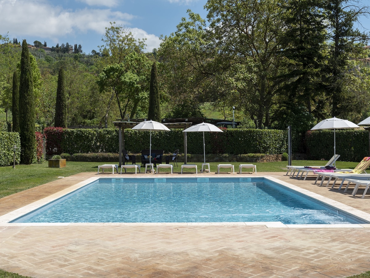 A serene shared swimming pool is surrounded by lush greenery and partially shaded by white umbrellas. Lounge chairs are arranged around the water, inviting relaxation under the clear sky. The pool is fenced for safety, and vibrant trees create a peaceful atmosphere.