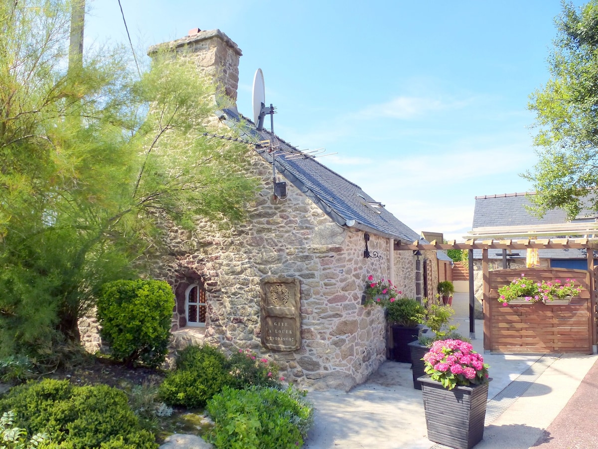 A charming stone cottage features a textured exterior accented by greenery and colorful flower pots. A small pathway leads to the entrance, where sunlight illuminates the surrounding plants. A satellite dish is mounted on the roof, indicating modern amenities amid rustic architecture.