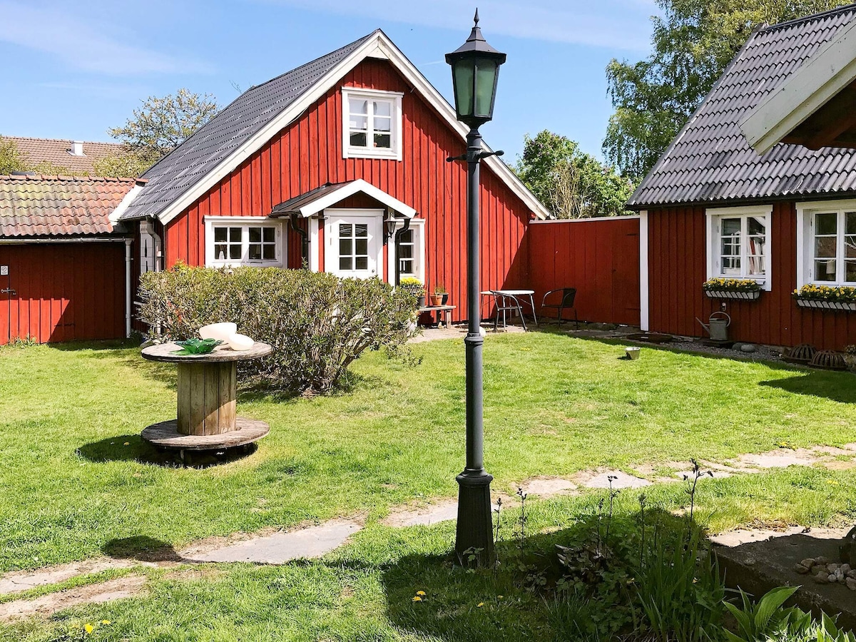 A charming red farmhouse is surrounded by greenery in the yard. A stone path leads through the grass, passing a wooden table and chairs set for outdoor seating. A decorative lamp post adds character to the inviting outdoor space.