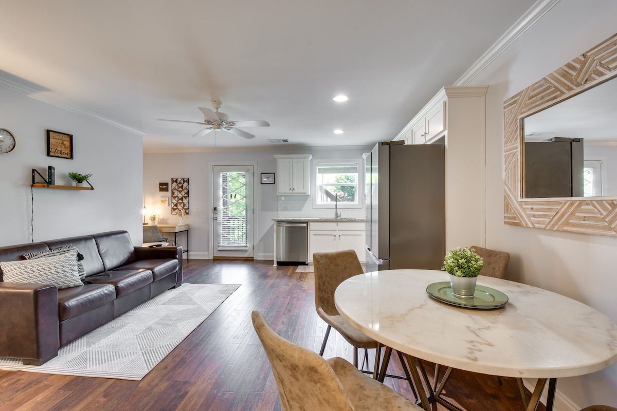 An inviting living space features a plush brown sofa and a round dining table with four chairs. Natural light fills the room through large windows, illuminating the polished wood floors. A mirrored accent piece adorns one wall, enhancing the open layout.