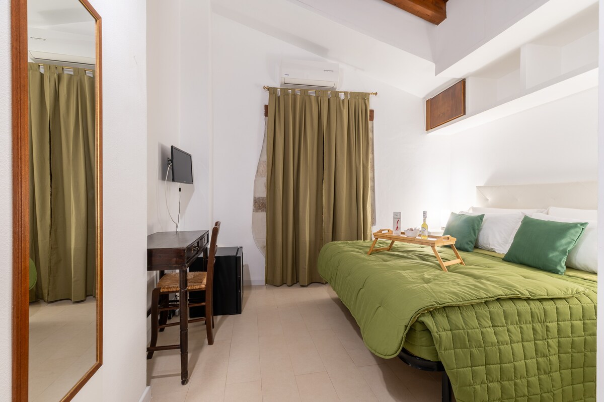 A bright room features a spacious bed dressed in a vibrant green coverlet. A simple desk and chair are positioned nearby, while a large mirror reflects the cozy space. Natural light is filtered through long, olive-green curtains, complementing the minimalistic décor.