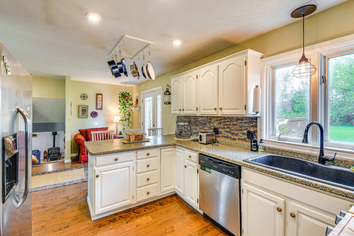 The kitchen features white cabinetry complemented by a stone backsplash. A stainless-steel refrigerator is positioned next to a dishwasher. Natural light enters through multiple windows, illuminating the granite countertops and a sink. A cozy seating area is visible in the background.