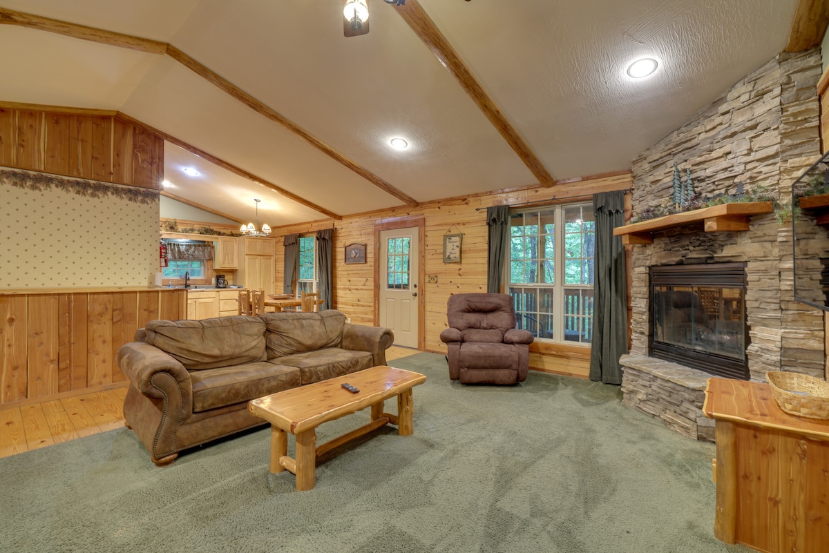 A spacious living area showcases soft seating arrangements, including a couch and recliner, arranged around a natural stone fireplace. Large windows allow ample light and views of the surrounding forest. Wooden beams and paneling create a rustic charm, complementing the carpeted floor.
